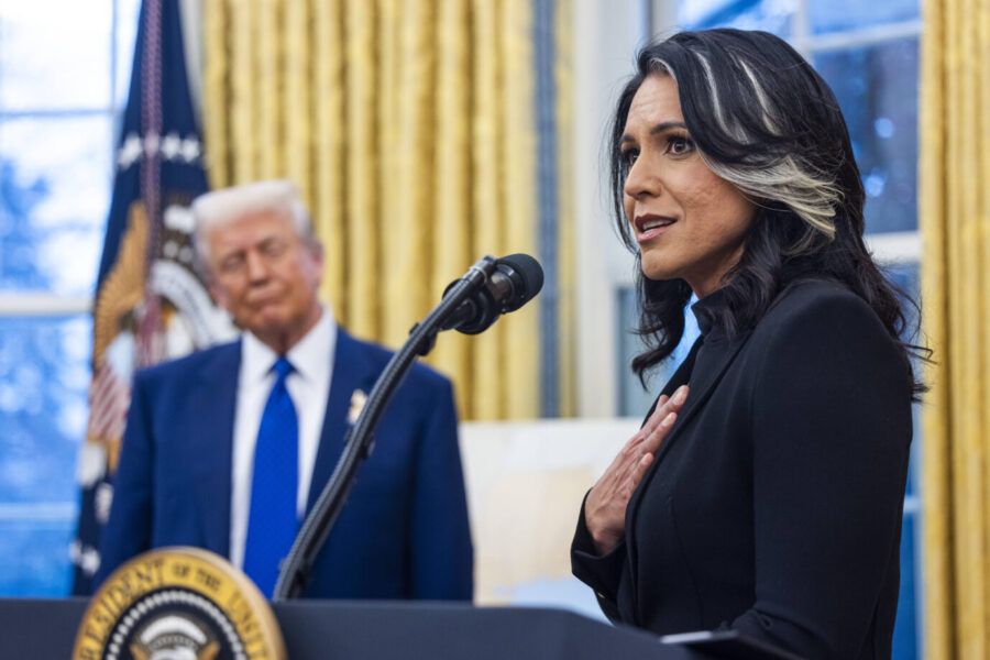 Tulsi Gabbard spricht mit der Hand auf der Brust vor einem Mikrofon im Weißen Haus, während Ex-Präsident Donald Trump im Hintergrund unscharf zu sehen ist und sie beobachtet; im Hintergrund goldene Vorhänge und das Präsidentensiegel.
