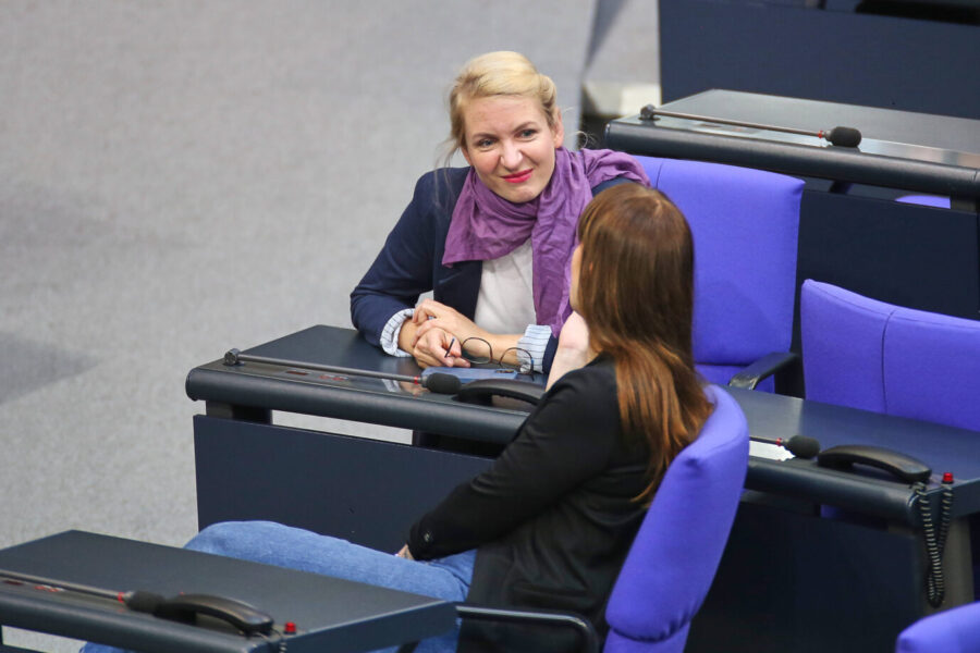 Zwei Frauen sitzen in einer Plenarsitzung auf lila Sesseln im Deutschen Bundestag. Eine von ihnen, mit blondem Haar und lila Schal, blickt lächelnd zur anderen. Vor ihnen liegen Mikrofone und Brillen auf den Tischen.