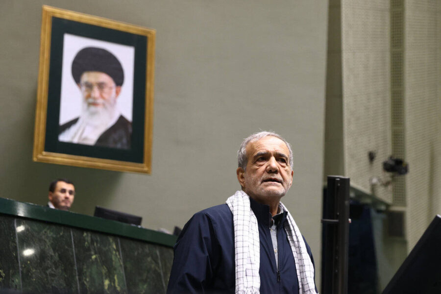Irans Präsident Masoud Pezeshkian steht in einem Sitzungssaal des Parlaments in Teheran. Er trägt eine dunkle Jacke und einen weiß karierten Schal über den Schultern. Im Hintergrund hängt ein großes Porträt von Ajatollah Ali Chamenei an der Wand. Neben Pezeshkian ist ein weiterer Mann hinter einem Pult zu sehen. Die Szene spielt sich am 16. Juni 2025 ab. Irans Präsident Masoud Pezeshkian bei einer Parlamentssitzung in Teheran – unter den Augen des Revolutionsführers. Foto: IMAGO / ZUMA Press Wire