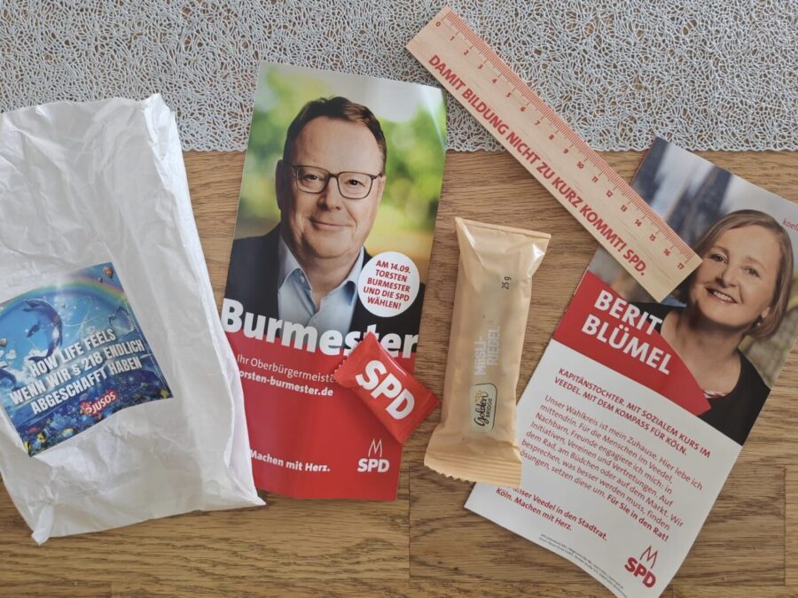 Auf einem Holztisch liegen Wahlkampfmaterialien: ein weißes Tütchen mit Juso-Aufkleber „How Life feels – Wenn wir Paragraph 218 endlich abgeschafft haben“, ein Flyer von Torsten Burmester, ein Flyer von Berit Blümel, ein Holzlineal mit SPD-Slogan, ein Müsliriegel und ein rotes Bonbon mit SPD-Logo. Inhalt der Juso-Tüten vor der Gesamtschule Rodenkirchen: Aufkleber zur Abschaffung des Paragraphen 218, Flyer der SPD-Kandidaten Torsten Burmester und Berit Blümel, Lineal, Müsliriegel und Bonbon mit SPD-Aufdruck. Foto: AfD Köln
