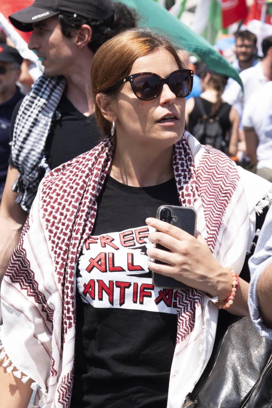 Die italienische Europaabgeordnete Ilaria Salis läuft während einer Demonstration in Rom am 21. Juni 2025 durch eine Menschenmenge. Sie trägt eine dunkle Sonnenbrille, eine rot-weiße Keffiyeh um die Schultern und ein schwarzes T-Shirt mit der Aufschrift „Free all Antifa“. In der Hand hält sie ein Smartphone, im Hintergrund sind weitere Teilnehmer mit Fahnen zu sehen. Salis ist Mitglied des Europäischen Parlaments und steht wegen mutmaßlicher Verbindungen zur linksextremen Hammerbande in der Kritik.Die italienische Abgeordnete Ilaria Salis bei einer Demonstration in Rom: Sie trägt eine Keffiyeh und ein T-Shirt mit der Aufschrift „Free all Antifa“. Foto: IMAGO / Avalon.red