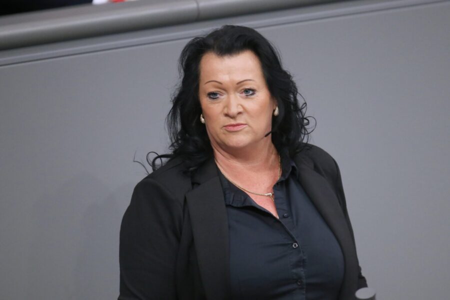 Eine Frau mit dunklen, lockigen Haaren steht in einem dunklen Blazer und einer schwarzen Bluse vor einer grauen Wand. Sie blickt leicht zur Seite und trägt goldene Ohrringe und eine Halskette. Die AfD-Abgeordnete Birgit Bessin am Rednerpult im Bundestag: „Luxusmigranten im Sozialsystem“. Foto: picture alliance / dts-Agentur | -