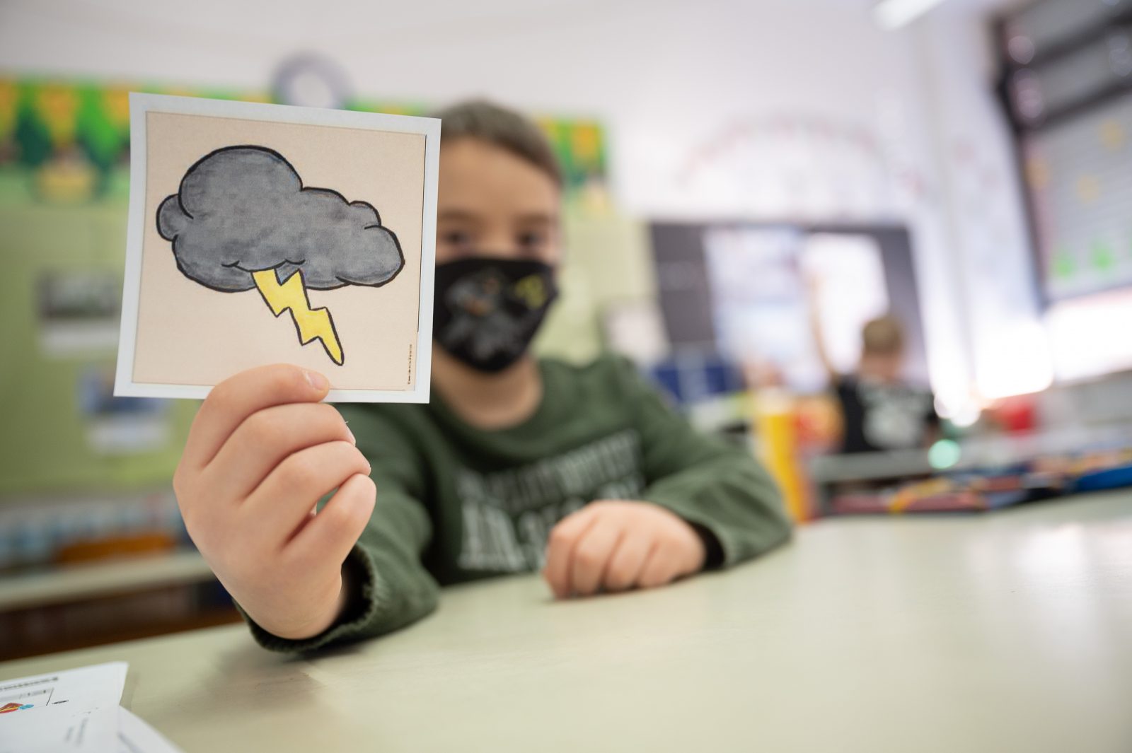 Ein Erstklässler hält einer Grundschule mit Wechselunterricht eine Karte mit einer Gewitterwolke hoch.