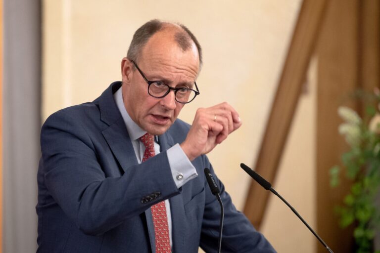 Friedrich Merz, Oppositionsführer und CDU-Parteivorsitzender, steht im dunkelblauen Anzug und mit roter Krawatte vor einem Mikrofon. Er gestikuliert mit seiner rechten Hand und sieht konzentriert aus.