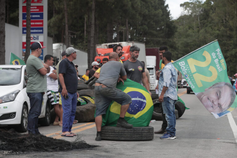Menschen stehen am Straßenrand. Sie tragen eine brasilianische Flagge und ein Banner mit dem Gesicht von Jair Bolsonaro.