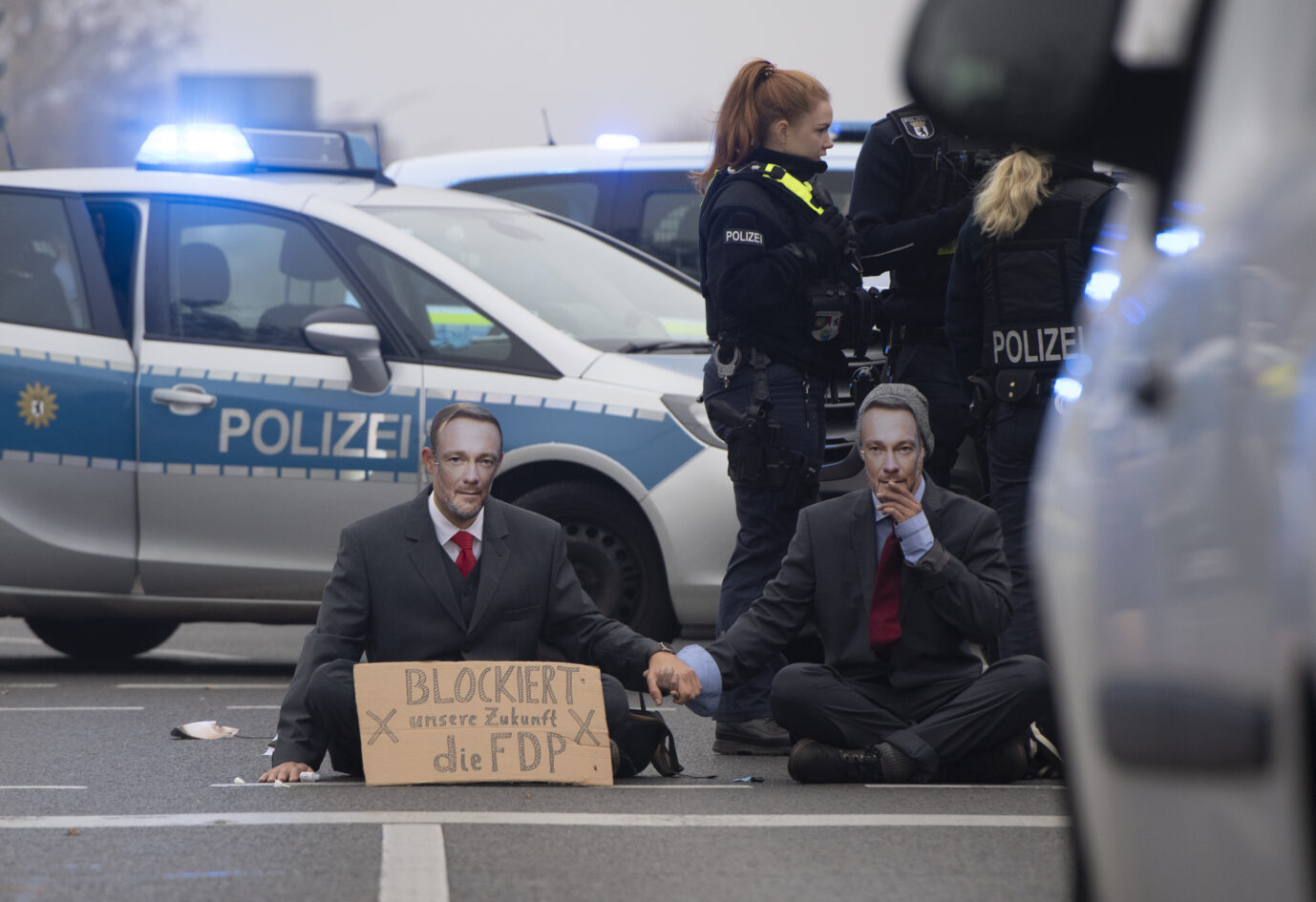 Zwei Mitglieder der "Letzten Generation" sitzen vor einem Polizeiauto auf der Straße. Sie tragen Anzüge und Masken mit dem Gesicht von FDP-Politiker Christian Lindner.