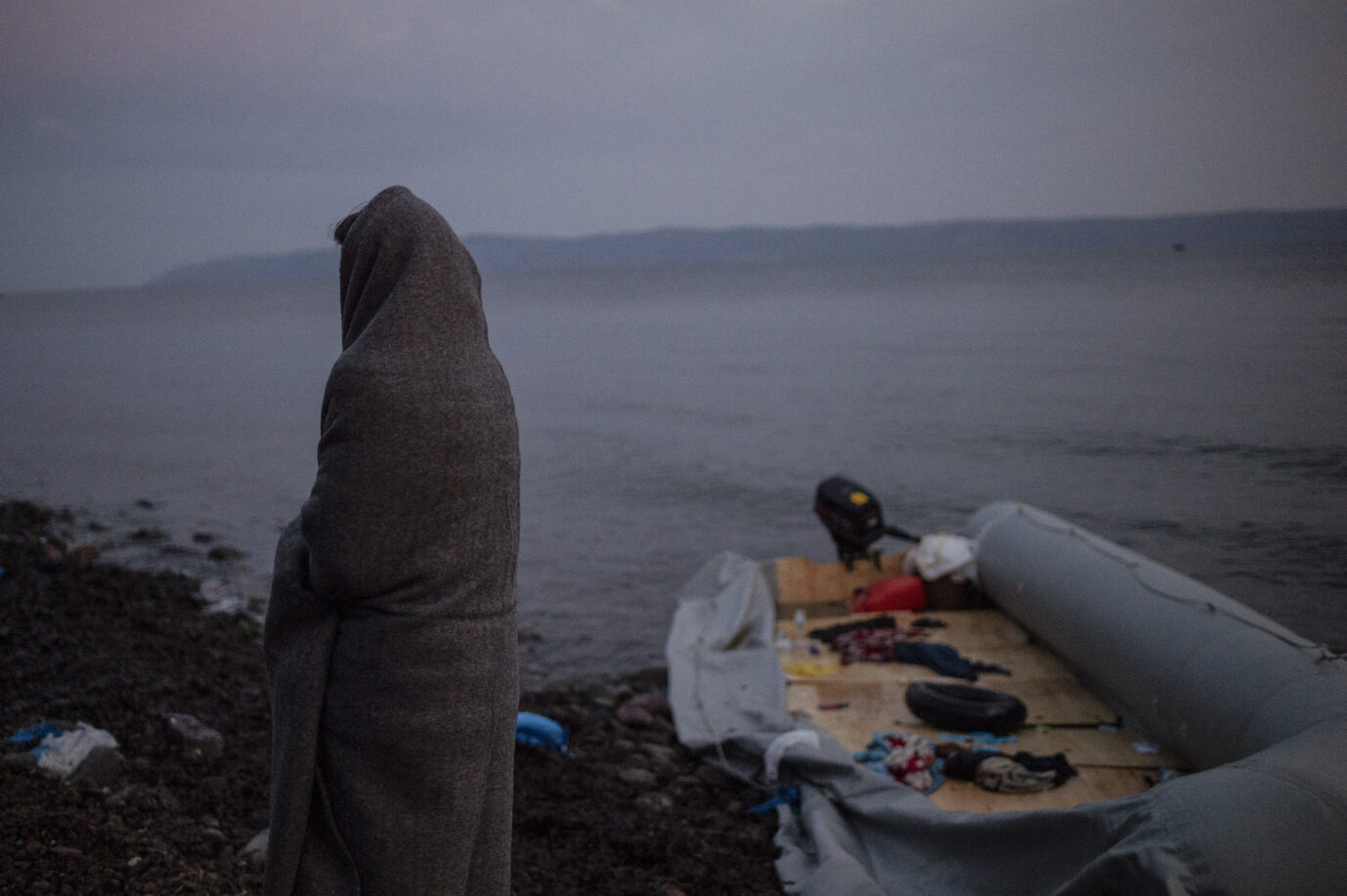 Eine Person ist von hinten sichtbar und in eine Decke eingewickelt an der Mittelmeerküste. Im Hintergrund ist ein Schlauchboot zu sehen. Illegale Migration ist zuletzt wieder angestiegen.