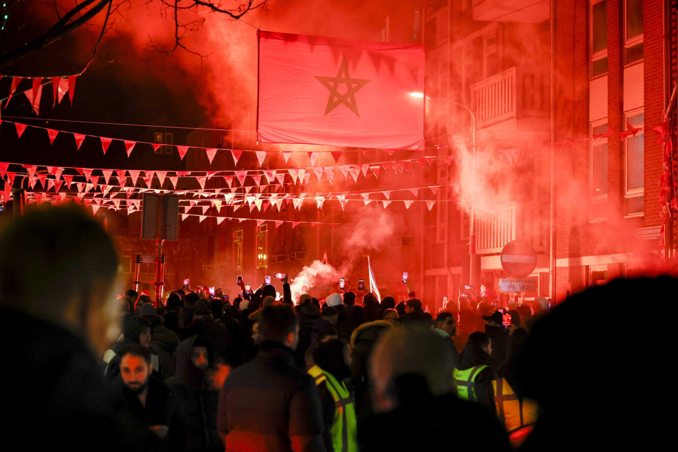 Eine dunkle Straße ist voller Menschen, rote Pyrotechnik leuchtet. Über der Straße hängt eine große Landesflagge von Marokko.