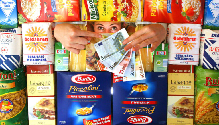 Das Bild zeigt eine Frau, die hinter einem Supermarktregal steht und eine Hand voll Geldscheine in die Kamera hält. Das Foto problematisiert die gestiegenen Lebensmittelpreise.