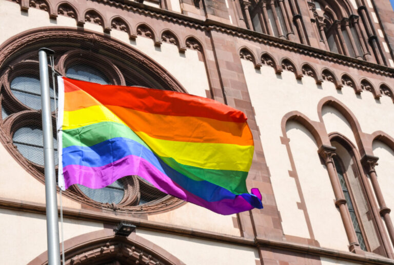 Eine Regenbogenflagge weht vor einer Kirche. In Kirchen wird gepredigt.