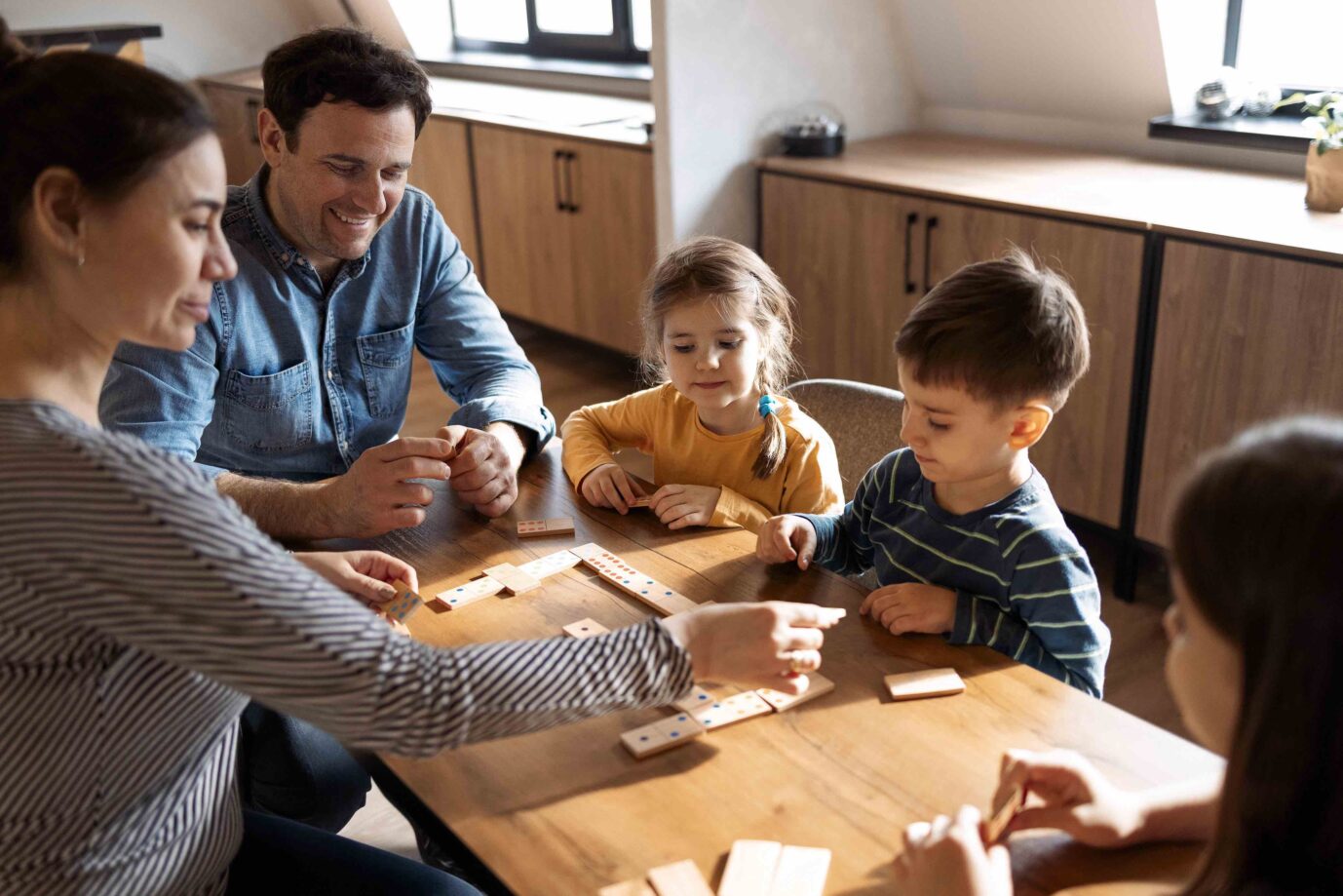 Mutter, Vater und Kinder spielen im Corona-Lockdown Domino, während sie im Wohnzimmer in der Nähe des Fensters sitzen. Die Familie verbringt Zeit miteinander.