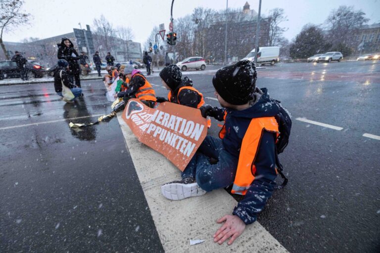 Mehrere Aktivisten der „Letzten Generation“ kleben sich für das Klima auf der Straße fest. Eine Tube Klebstoff liegt neben einem jungen Mann.