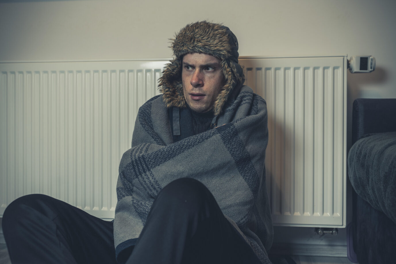 junger Mann sitzt frierend warm angezogen in seiner Wohnung vor der Heizung mit Wintermütze und Decke bedeckt. Symbolbild defekte Heizung und kalte Wohnung || Modellfreigabe vorhanden