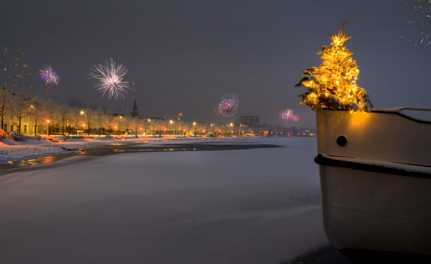 Weihnachtsbaum steht am Rande des Bugs eines Bootes und Feuerschwaden im Hintergrund. Silvesterzeit auf der Schlei. Feuerwerk an Silvester. Schneebedeckte Promenade im Winter in Schleswig.