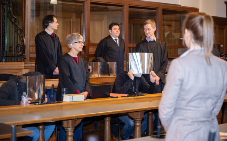 Vier Angeklagte sitzen vor Beginn der Verhandlung im Gerichtssaal im Kriminalgericht Moabit in Begleitung ihrer Anwälte Regina Götz (vorne l-r), Einar Aufurth, Marten Mittelstädt (hinten l-r) und Michael Lippa und verdecken dabei ihre Gesichter mit Pappe. Fast vier Jahre nach dem Angriff auf ein ZDF-Team am Rande einer Demonstration gegen die staatlichen Corona-Maßnahmen in Berlin beginnt der Prozess gegen drei Männer und eine Frau aus Berlin und Baden-Württemberg.