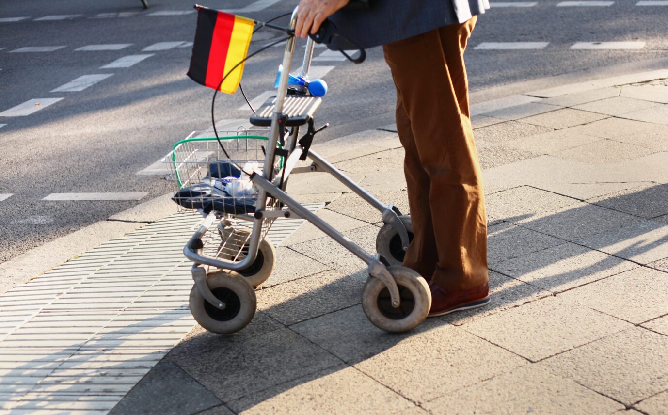 Ein Mann steht mit seinem Rollator, an dem ein Deutschlandfähnchen hängt, an einer Straße. Aufgrund der niedrigen Geburtenrate überaltert Deutschland zunehmend