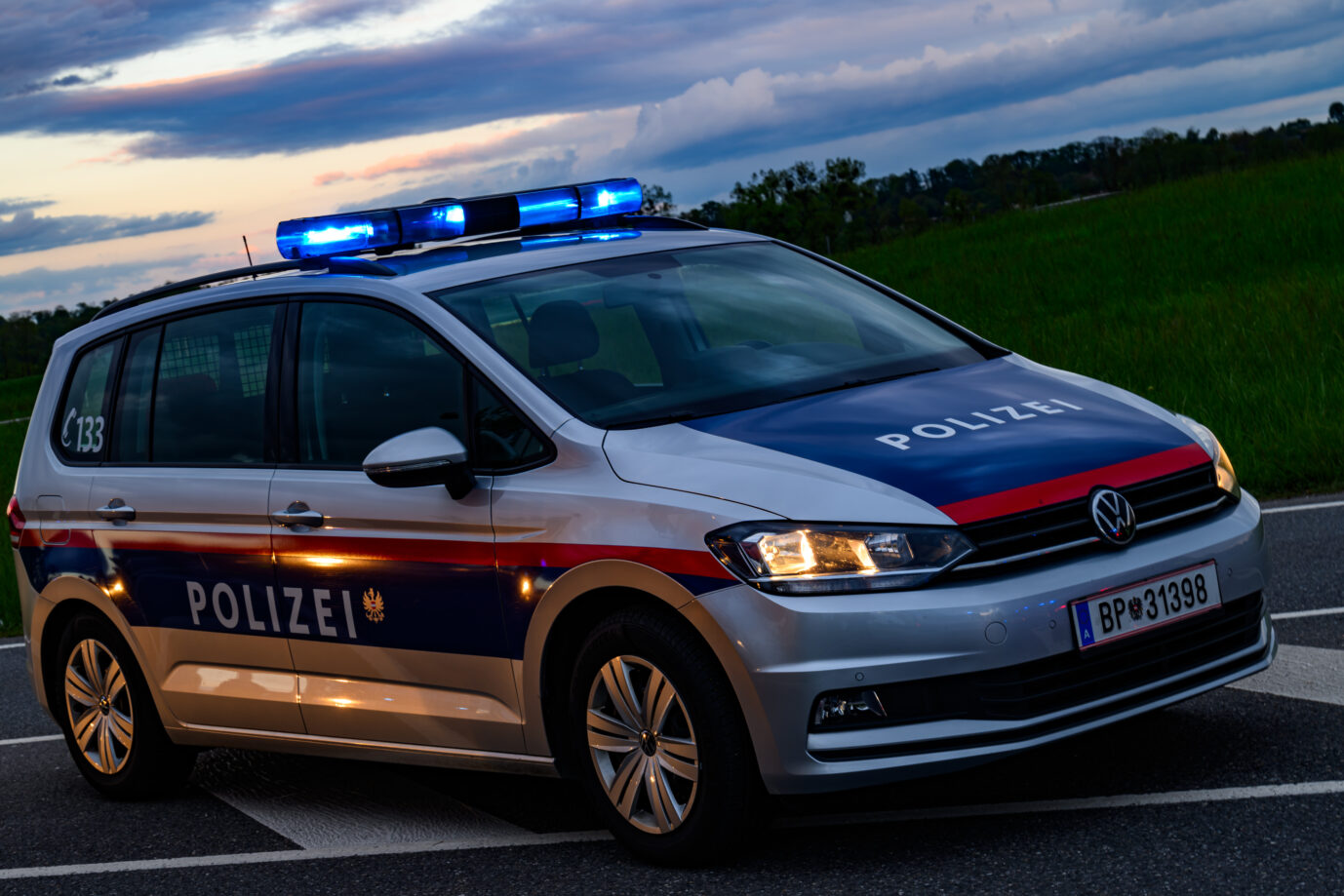 Ein Polizeifahrzeug der österreichischen Polizei steht einsatzbereit an der Straßenseite. Die Dämmerung setzt ein, während die Blaulichter des Fahrzeugs hell in den frühen Abendhimmel strahlen und die Umgebung in ein weiches Licht tauchen. In Niederösterreich hat ein Afghane schwere Straftaten begangen. - 20240421_PD18121