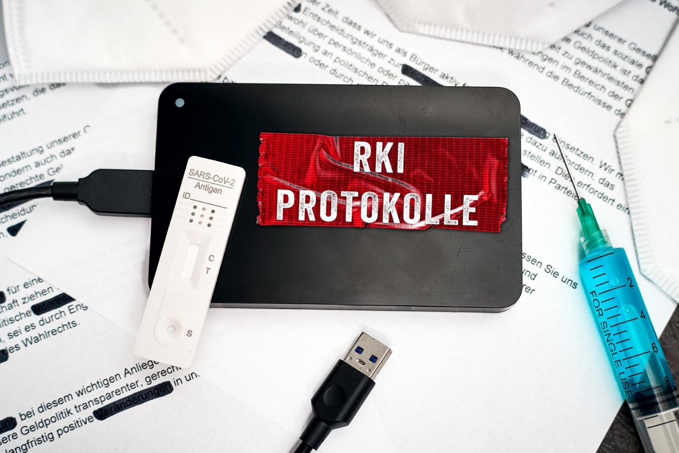 Symbolbild Robert Koch Institut Dokumente zu Covid-19. Eine Festplatte mit Aufschrift RKI Protokolle liegt auf geschwärzten Dokumenten zusammen mit FFP2-Masken, Spritze und Schnelltest. FOTOMONTAGE