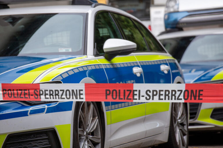 Ein deutsches Polizeiauto steht hinter einer rot-weißen Absperrung – möglicherweise ein abgesperrter Tatort nach einem Messerangriff oder einem Ehrenmord