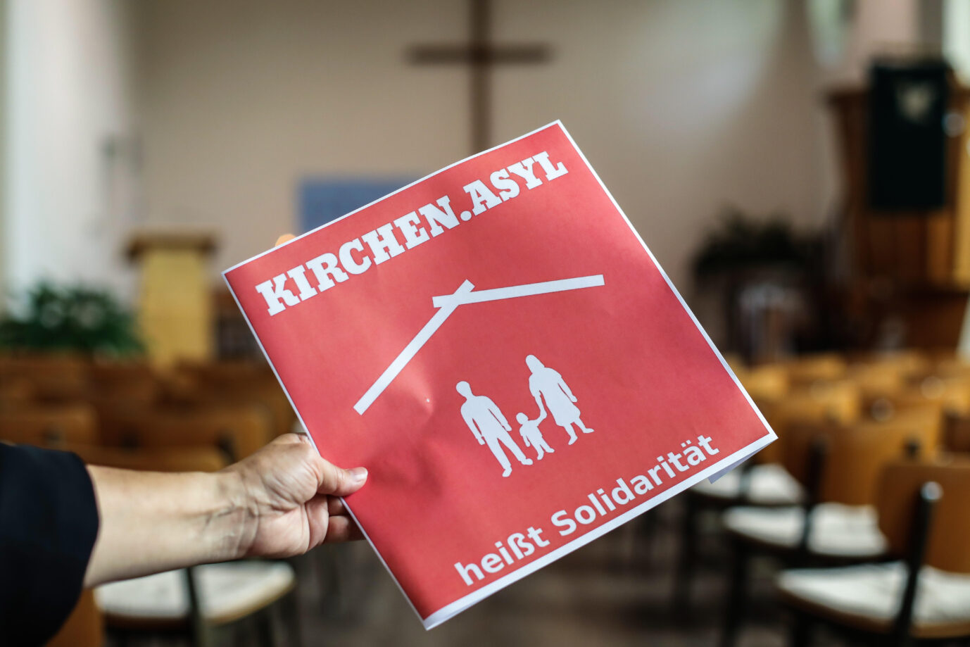 Ein rotes Schild mit weißem Rand und drei weißen Vater-Mutter-Kind-Figuren, die häufig im Zusammenhang mit dem Thema Asyl verwendet werden und der Aufschrift Ein Schild mit der Aufschrift „Kirchenasyl heißt Solidarität“