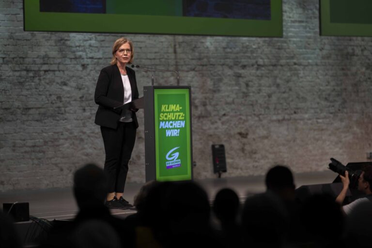 Österreichischs Umweltministerin Leonore Gewessler (Grüne) trägt Sakko und Bluse und steht vor einem grünen Rednerpult mit der Aufschrift: "Klimaschutz. Machen wir"