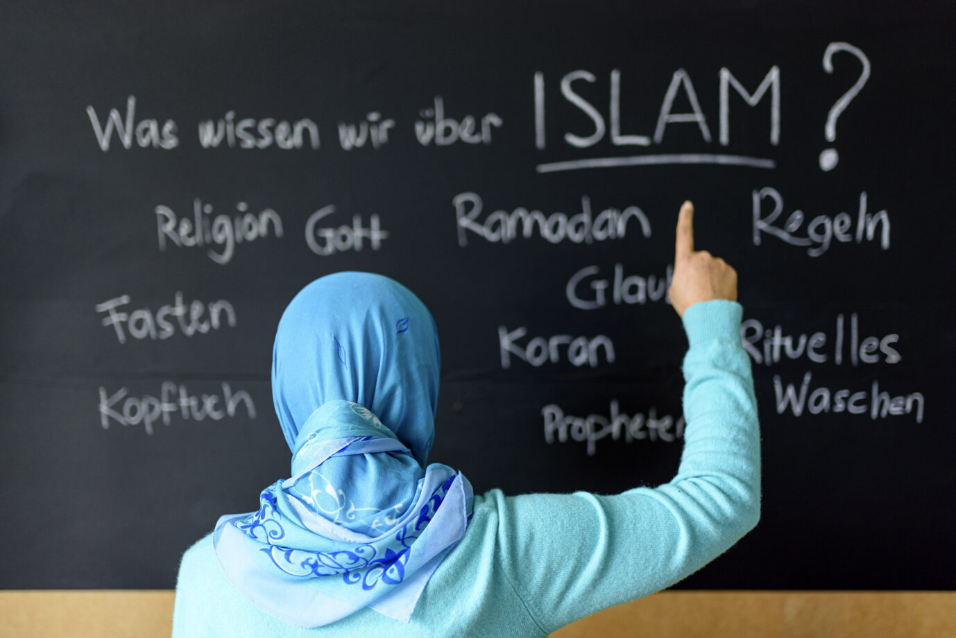 Ein junges Mädchen mit einem hellblauen Kopftuch schreibt mit einem Stück Kreide Begriffe wie „Islam“, „Gott“ und „Ramadan“ an eine Schultafel – wurde sie etwa von der Scharia-Polizei angestiftet?