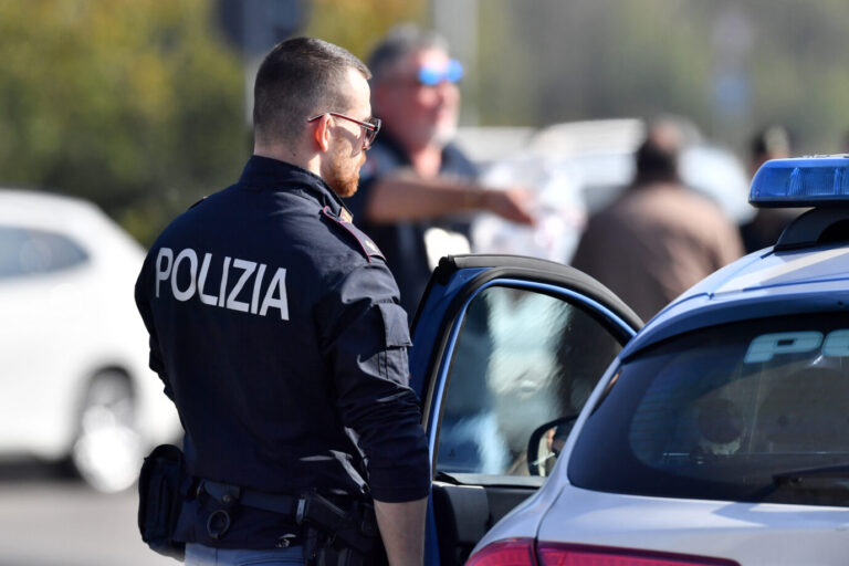 Ein Mann in der Uniform der italienischen Polizei steht vor einem Polizeiauto, im Hintergrund ist verschwommen der Tatort zu sehen. So könnte eine Szene anläßlich der Vergewaltigung in Turin aussehen