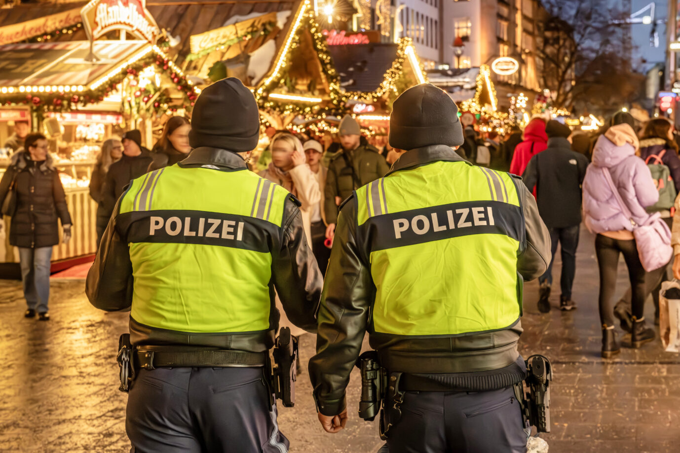 Zwei Polizeibeamte in gelben Warnjacken patrouillieren auf einem Weihnachtsmarkt in München. Im Hintergrund sind die bunten Lichter eines Standes zu sehen