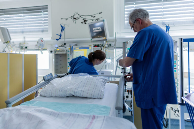 Zwei Ärzte oder Pflegekräfte stehen an einem Krankenhausbett, arbeiten daran herum und tragen blaue Kittel. Das Bild symbolisiert die Krankenhausreform von Karl Lauterbach