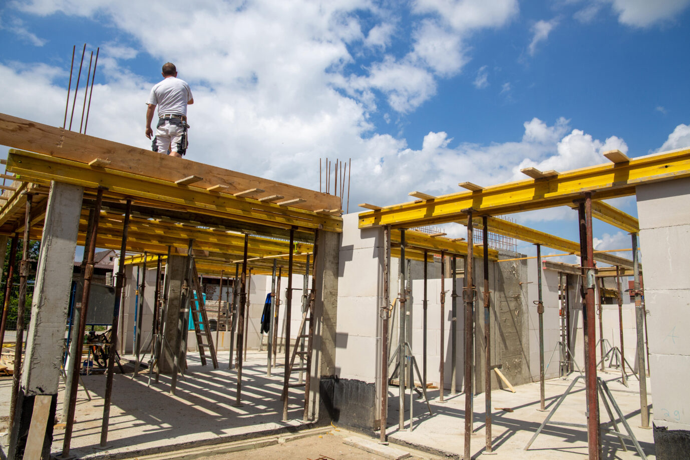 Auf einer Baustelle sind Metallstäbe und Zementblöcke zu sehen. Dazwischen steht ein Maurer oder Arbeiter in Arbeitskleidung. Hier wird eine Wohnung gebaut