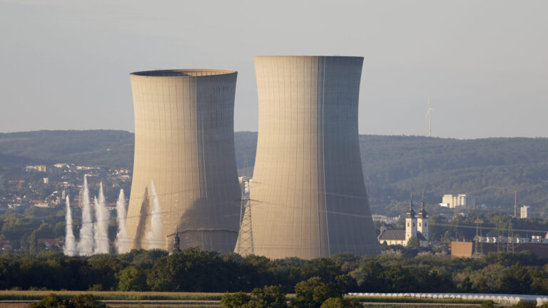 Zwei graue Kühltürme eines Kernkraftwerks in Grafenrheinfeld knicken ein und stoßen weißen Rauch aus – sie werden gesprengt. Zugunsten von Klimaschutz und Energiewende