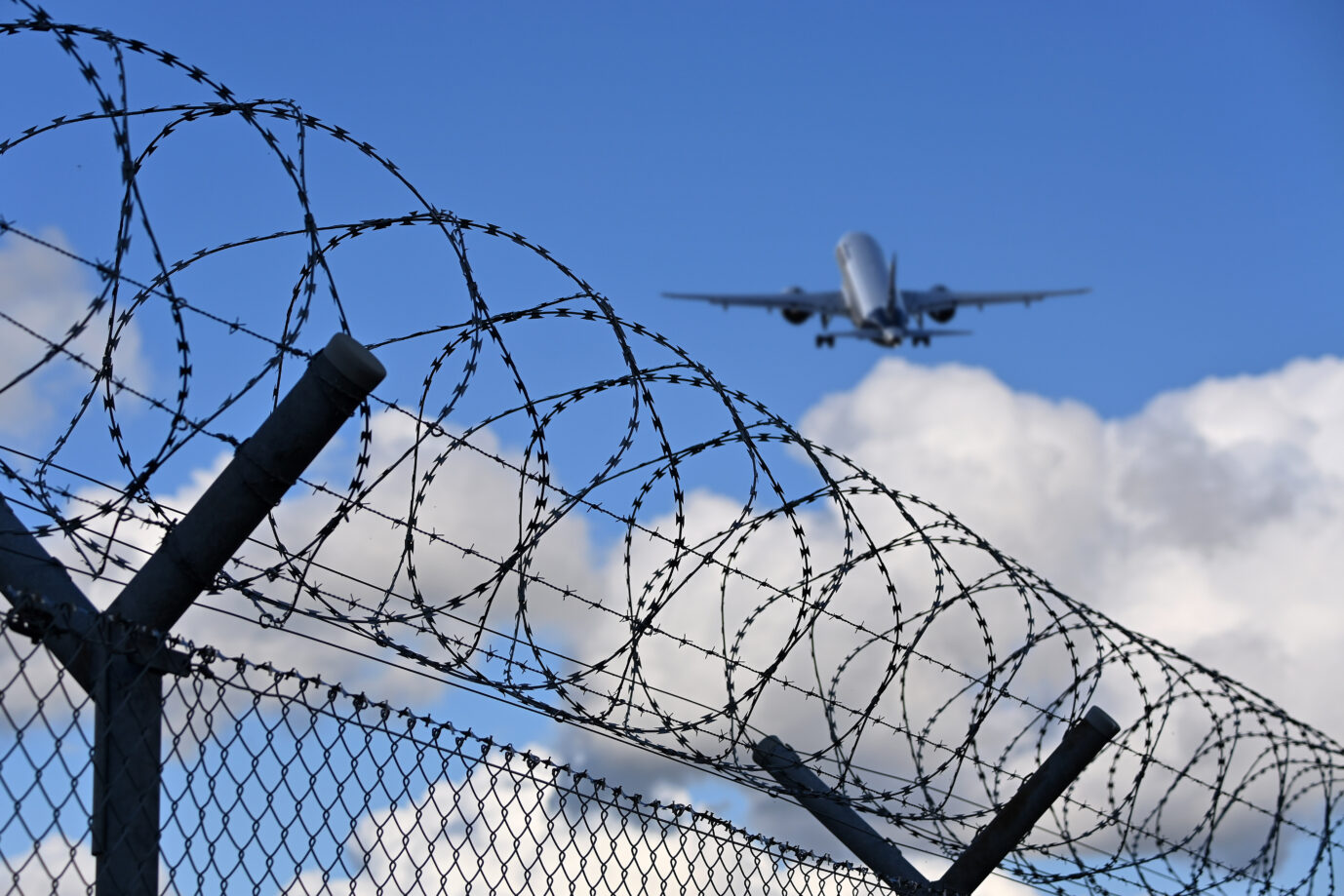 In einem blauen bewölkten Himmel hebt ein Passagierflugzeug ab, davor ist Stacheldraht zu sehen