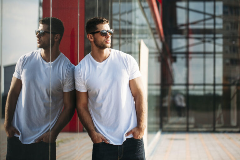 Ein Mann mit Sonnenbrille und Dreitagebart trägt ein schlichtes weißes T-Shirt und lehnt lässig an einer Hauswand