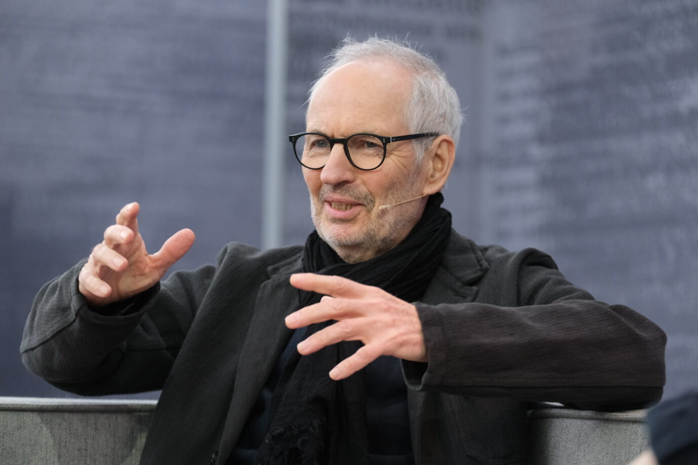 Schriftsteller Eugen Ruge, gekleidet in einen schwarzen Pullover, mit schwarzem Schal und schwarzer Hornbrille, gestikuliert mit den Händen – das Foto entstand während einer Rede auf der Leipziger Buchmesse, 2023