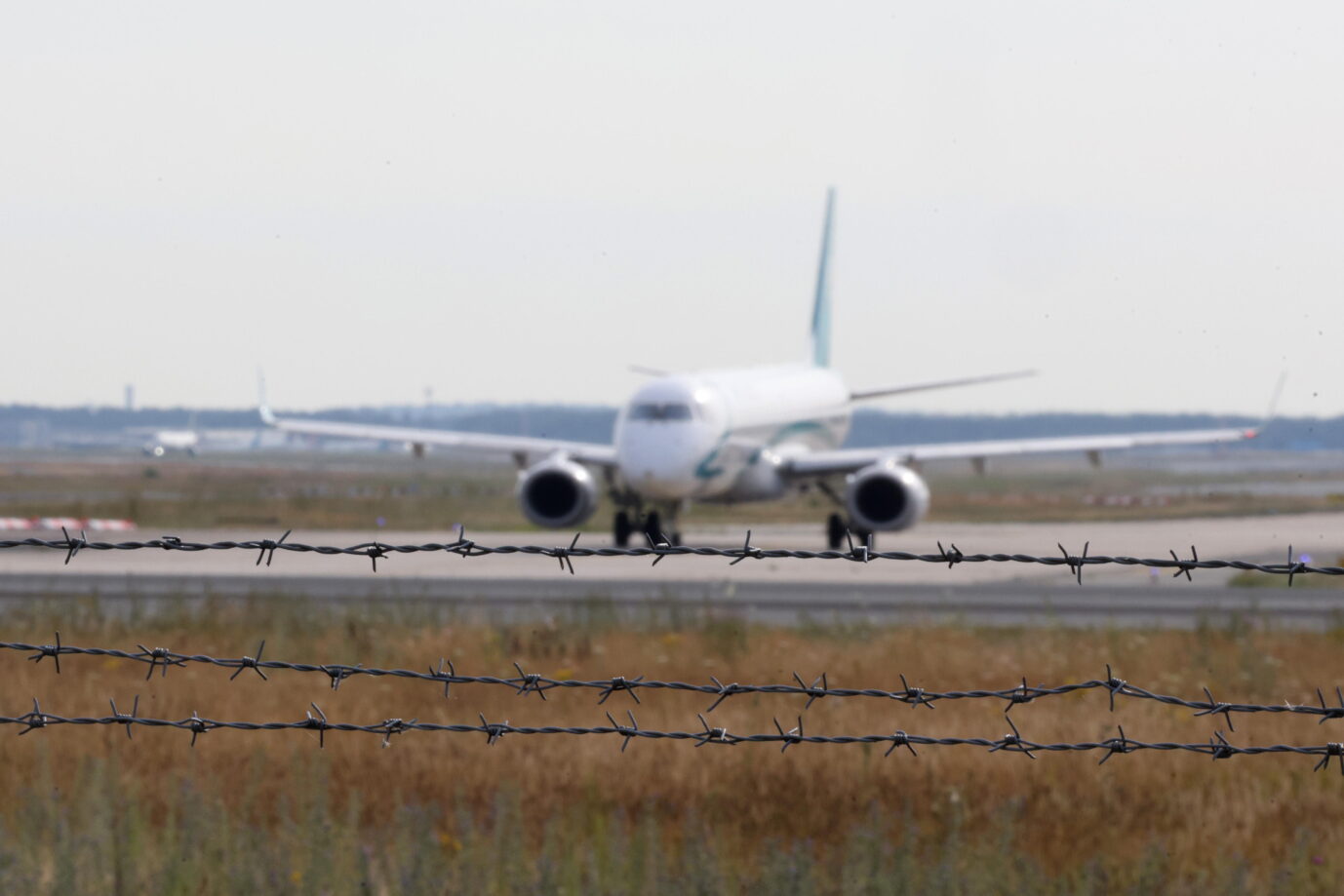 Auf dem Rollfeld eines Flughafens steht ein Flugzeug, im Vordergrund ist Stacheldraht zu sehen- Das Flugzeug symbolisiert Abschiebungen