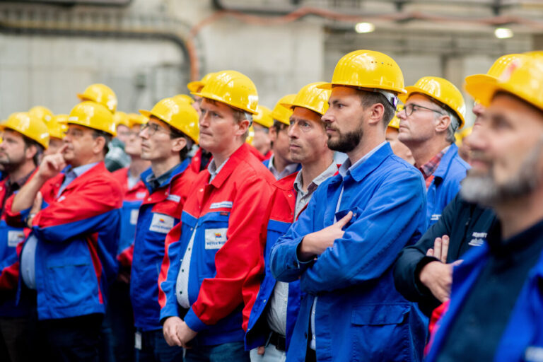 Arbeiter mit gelben Schutzhelmen und blau-roten Westen stehen während einer Betriebsversammlung in einer Werfthalle des Unternehmens Meyer Werft. Auch die Werft muß massiven Jobabbau betreiben