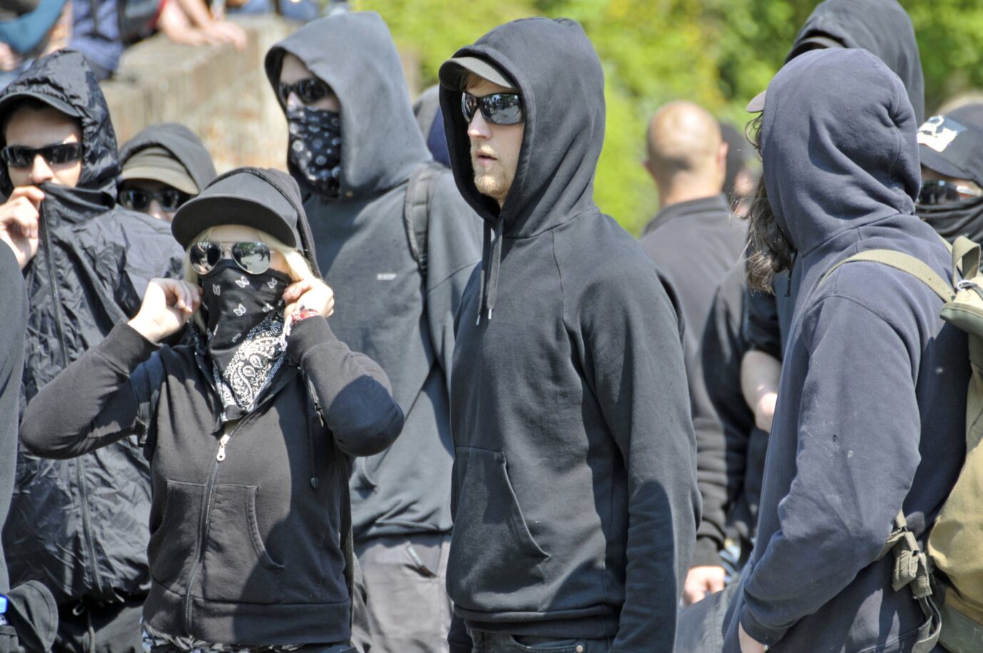 Vermummte und in schwarz gekleidete Jugendliche stehen auf einer Demonstration herum. Es handelt sich um linksextreme Mitglieder der Antifa