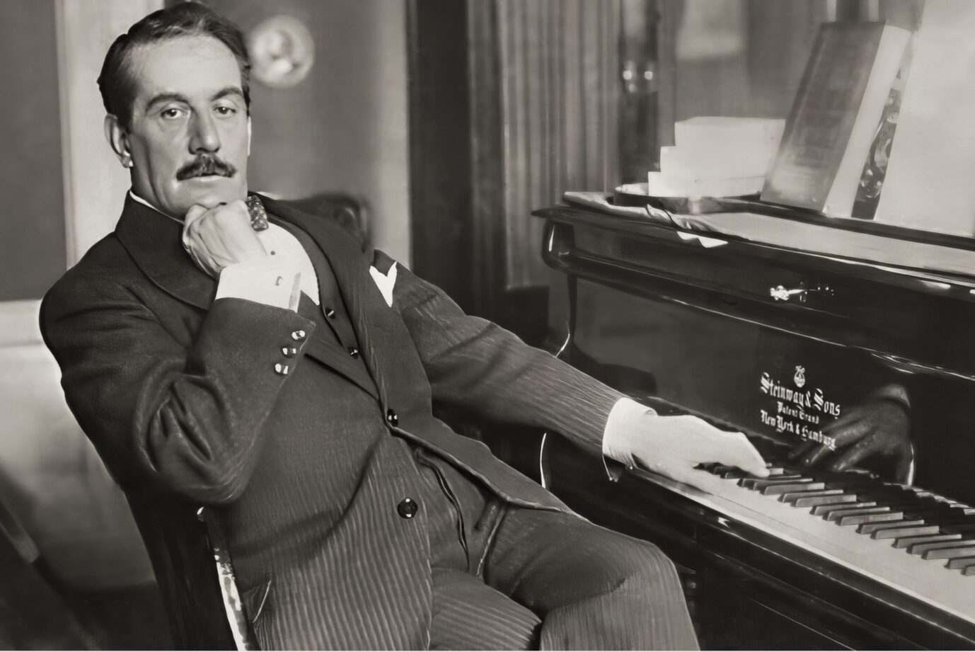 Das alte Schwarzweißbild zeigt den italienischen Musikkomponisten Giacomo Puccini (1858–1924), an einem Klavier sitzend und die rechte Hand aufs Kinn gestützt