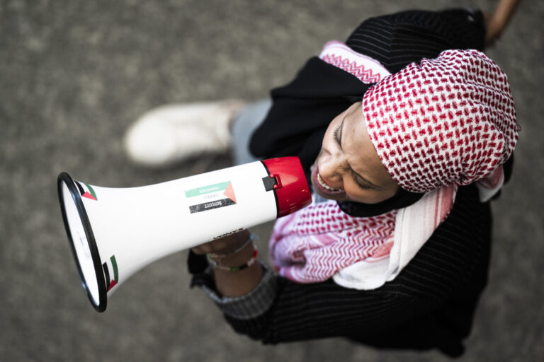 Eine junge Frau arabischen oder nordafrikanischen Aussehen trägt ein sogenanntes Palästinensertuch, ein Kufiya, und brüllt in ein Megafon. Es ist eine Pro-Palästina-Demonstrantin