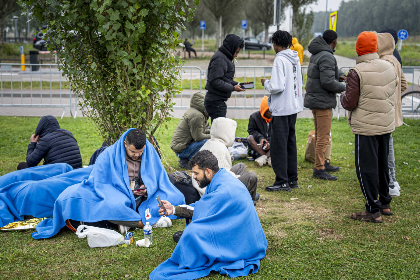 Auf einer Wiese sitzen junge Asylbewerber – arabische und afrikanische Männer – und warten, daß das Antragszentrum sie reinläßt – dank der neuen Schuldenbremse werden die Bundesländer demnächst mehr Geld bzw. Schulden für sie ausgeben