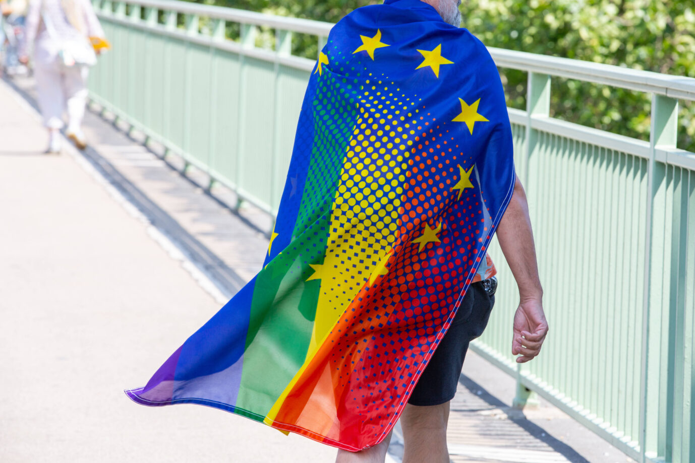 Auf einer Brücke steht ein Mann der sich eine Flagge umgehängt hat – die Flagge zeigt eine Hybridsymbolik aus EU- und Regenbogenlogo – Symbolbild für die Gender-Agenda der EU, die diese jetzt auch Ungarn aufzwingen will