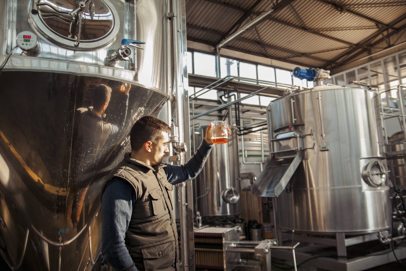 Ein Arbeiter hält in einer Brauerei ein Glas Bier hoch, im Hintergrund sind schwere Maschinen zu sehen