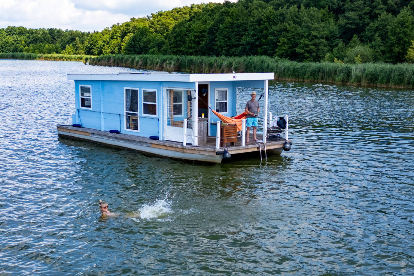 Ein blau-weißes Hausboot schwimmt auf dem Wasser, eine Frau geht schwimmen und der Mann schaut zu