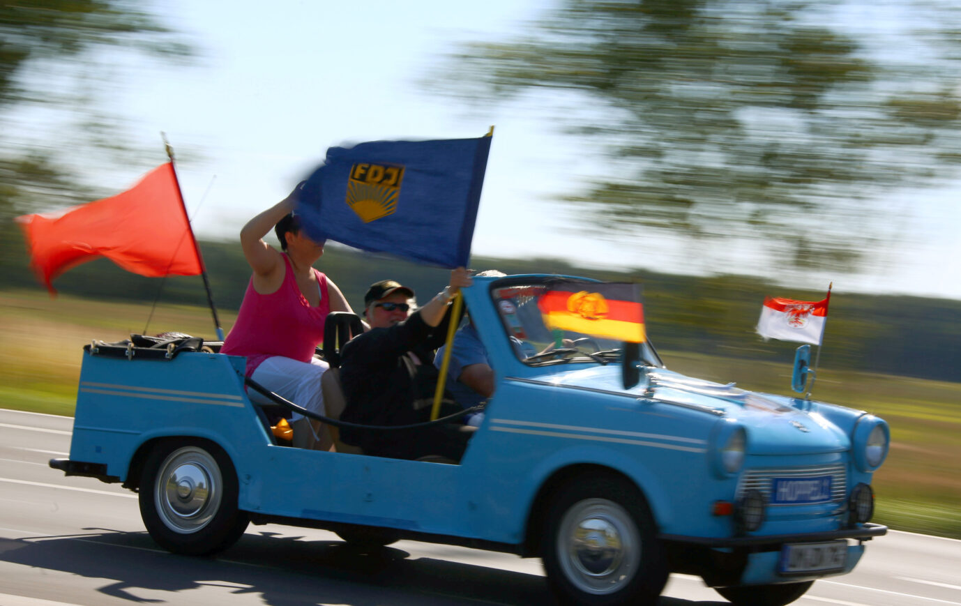 Auf einem blauen getunten Trabbi sitzen mehrere Personen und zeigen DDR-Fahnen – Symbolbild für die klischeehafte Medienberichterstattung über Ostdeutsche und Ostdeutschland