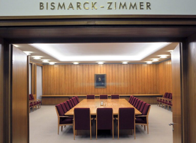 Blick durch eine geöffnete Holztür in einen Konferenzraum mit der Aufschrift ‚Bismarck-Zimmer‘ über dem Eingang. Im Raum steht ein großer, rechteckiger Holztisch mit 18 gepolsterten Stühlen in dunklem Rot. Die Wände sind holzvertäfelt, an der Stirnseite hängt ein gerahmtes Porträt von Otto von Bismarck. Die Beleuchtung ist in die Decke eingelassen und gleichmäßig verteilt. Der Raum wirkt aufgeräumt und traditionell eingerichtet. Das Bismarck-Zimmer im Auswärtigen Amt war zu DDR-Zeiten der Politbürosaal. Statt Bismarck hingen hier Bilder von Marx und Lenin. Aufnahme vom 27.11.2008. Der traditionsreiche Saal mit holzvertäfelten Wänden: Wird der Raum wieder „Bismarck-Zimmer“ heißen? Foto: picture-alliance/ dpa | Markus C. Hurek