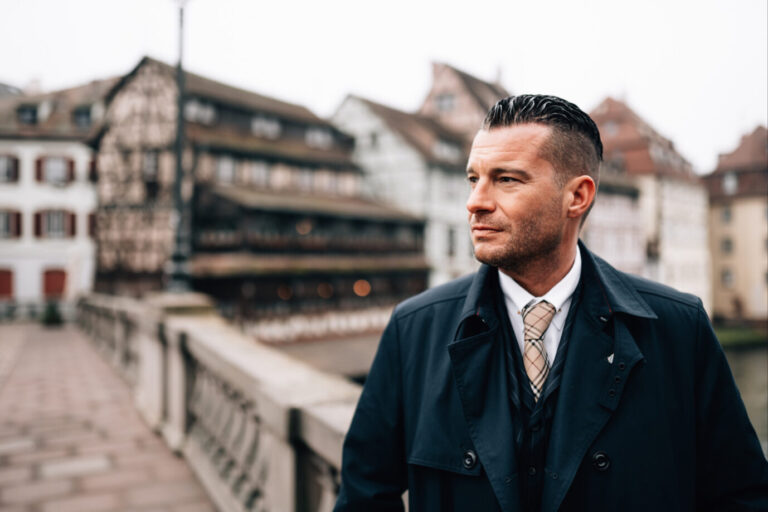 Ein Mann im Anzug mit gestreifter Krawatte und dunklem Mantel steht auf einer steinernen Brücke in einer historischen Altstadt mit Fachwerkhäusern. Er blickt ernst nach rechts. Der Hintergrund ist leicht unscharf. Sein Name ist David Bendels.