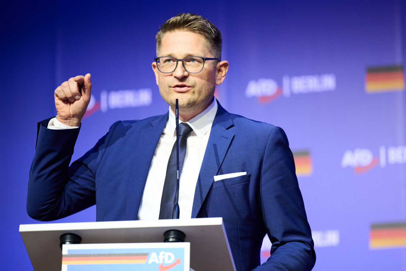 Der Brandenburger AfD-Landesvorsitzende René Springer steht vor einem blauen Hintergrund auf dem das AfD-Logo und die Deutschlandflagge prangt