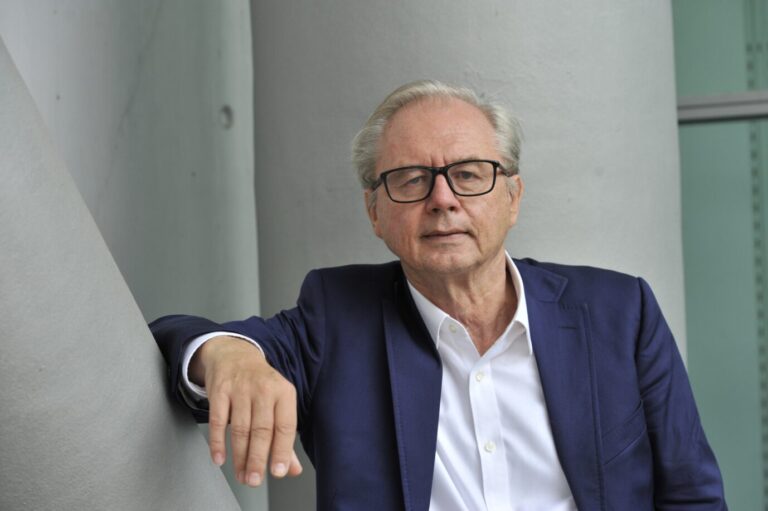 Der Journalist Wolfgang Herles trägt Jackett und ein weißes Hemd und lehnt sich lässig gegen einen Betonpfeiler