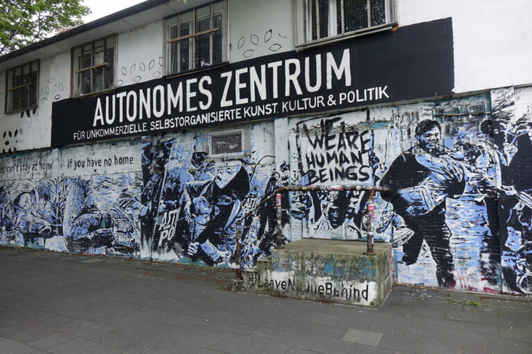 Außenansicht eines autonomen Zentrums mit großem Schriftzug „AUTONOMES ZENTRUM – Für unkommerzielle, selbstorganisierte Kunst, Kultur & Politik“ über dem Eingang. Die Fassade ist mit politischen Wandbildern und Graffiti bedeckt, darunter Parolen wie „You can't stay at home if you have no home“, „ANTI POL“ und „WE ARE HUMAN BEINGS“. Auf der Mauer vor dem Gebäude steht „#LeaveNoOneBehind“. Die Fenster sind vergittert, der Gesamteindruck wirkt alternativ und aktivistisch. Das „Autonome Zentrum“ in Kölns Luxemburger Straße: Die Stadt finanziert den Umzug. Foto: IMAGO / Steinach