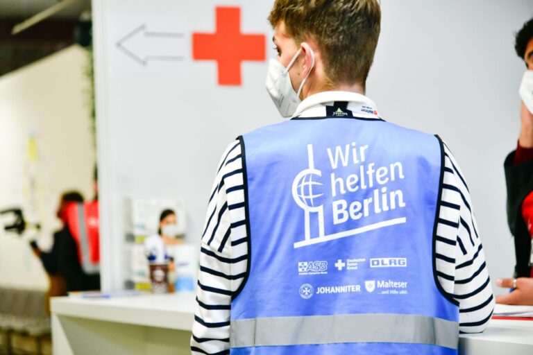Ein Helfer in der Asylunterkunft Berlin-Tegel trägt eine Maske und eine Weste mit der Aufschrift "Wir helfen Berlin.